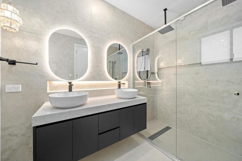 Floating black double vanity with backlit oval mirrors beside a frameless glass walk-in shower.